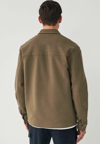Chaqueta de camisa verde oliva hecha de tejido texturizado, con un cuello de solapa, mangas largas y detalles de botones en los puños.