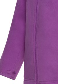 Manche de veste en polaire violette et partie avant avec fermeture éclair visible et petite boucle en tissu près du poignet.