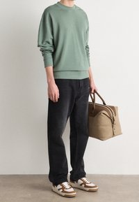 Persona che indossa una felpa verde salvia, jeans neri e sneakers bianco-marroni, con in mano una borsa da viaggio beige e marrone.