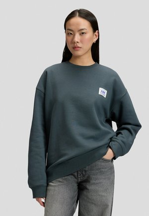 Frau mit glattem, dunklem Haar trägt ein dunkel-türkisfarbenes Lee-Sweatshirt und graue Jeans und steht mit einer Hand in der Tasche vor einem schlichten Hintergrund.