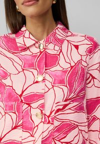 Veste florale en rose et rouge avec un tissu texturé. Dotée d'un col, de quatre boutons et de deux poches avant.