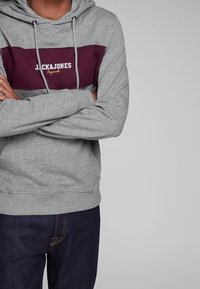 Sudadera gris con una franja granate que presenta el logo "JACK & JONES" en blanco. Cuerdas de ajuste y un bolsillo canguro, dobladillo y puños acanalados.