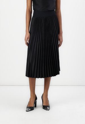 Persona llevando una falda midi plisada negra, camiseta negra sin mangas y tacones altos negros con punteras metálicas.
