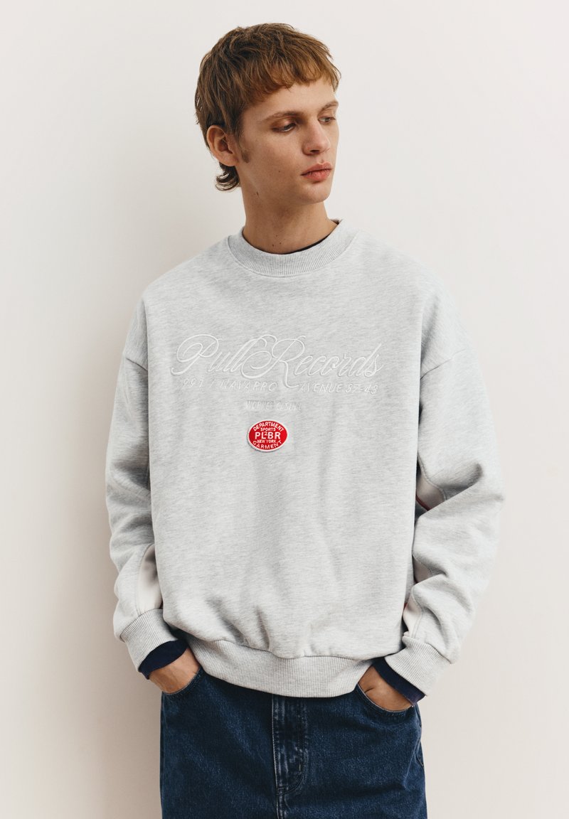 Grauer Fleece-Sweatshirt mit Rundhalsausschnitt, mit dem weißen Aufnäher "Pull Records" und einem roten Etikett auf der Vorderseite. Klassische Passform.