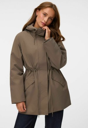 Femme portant une parka taupe à capuche avec des cordons à la taille et des poches à rabat, debout sur un fond clair uni.