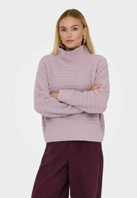 Un pull à col roulé lilas avec un motif en tricot texturé, des poignets et un ourlet côtelés, offrant une coupe décontractée. Assorti à un pantalon bordeaux foncé.