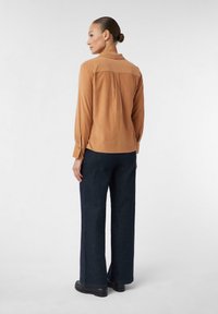 Camicia marrone a maniche lunghe con colletto, schiena pieghettata e polsini abbottonati, abbinata a pantaloni larghi blu scuro e calzature nere.