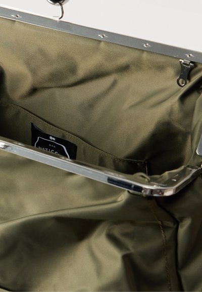 Ανοιχτό χακί υφασμάτινο τσάντα με μεταλλικό πλαίσιο και κούμπωμα, που παρουσιάζει εσωτερική θήκη με μαύρη ετικέτα.