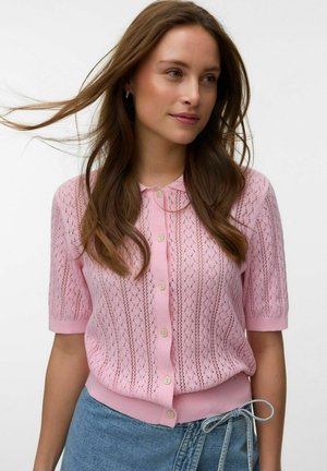 Young woman wearing a pink short-sleeve knit cardigan with buttons, paired with light blue jeans tied with a string belt, hair flowing.