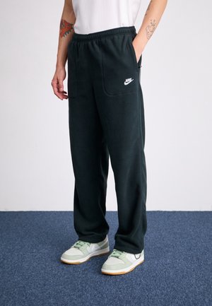 Mężczyzna w czarnych spodniach dresowych Nike, białej koszulce i jasnozielono-białych trampkach stoi na niebieskim dywanie na tle białej ściany.