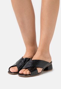 Mules noirs à bout ouvert, avec un design de lanières croisées. Fabriqués en simili cuir texturé avec un motif crocodile et un petit talon carré.