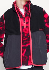 Giacca in pile nera e rossa con chiusura a zip e colletto alto. Presenta tasche a contrasto, un design a motivo e dettagli in neon rosa.