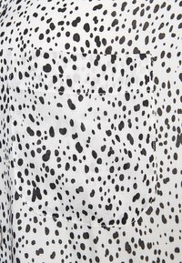 Tissu blanc avec des taches noires irrégulières et une poche carrée cousue se fondant dans le motif tacheté.