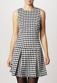 Mustaa ja valkoista houndstooth-kuosisilla sleeveless-mekolla, jossa on istuva yläosa ja liehukehame. Sileä kangas, polven ylle ulottuva pituus.