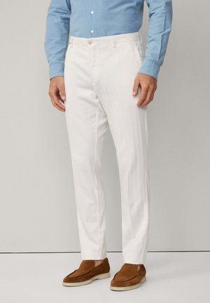 Uomo che indossa pantaloni bianchi su misura, camicia azzurra con bottoni e mocassini in pelle scamosciata marrone, in piedi contro uno sfondo chiaro uniforme.