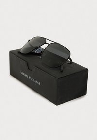 Zwarte metalen zonnebril met donkere lenzen, rechthoekige vorm en rubberen oordoppen, rustend op een zwarte doos met het label "Armani Exchange."