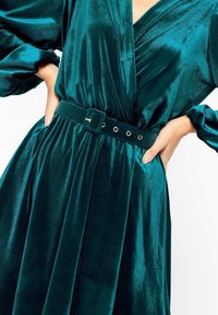 Teal sammet klänning med djup V-urringning, långa ärmar och midja med bälte. Bältet har fyrkantig spänne och silverögon. Mjuk, glänsande textur.