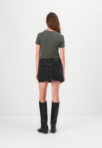T-shirt verde oliva a maniche corte, mini gonna in denim nera, stivali neri con tacco al ginocchio e sfondo bianco semplice.