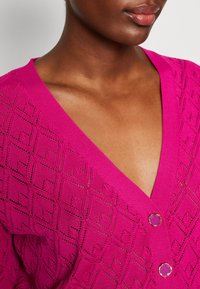 Cárdigan de punto magenta con un patrón de diamantes, que presenta un escote en V profundo y dos botones redondos decorativos en la parte delantera.