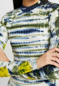 Haut à manches longues présentant un motif tie-dye aux nuances de bleu, vert et jaune. Le tissu a une texture lisse avec un col côtelé.