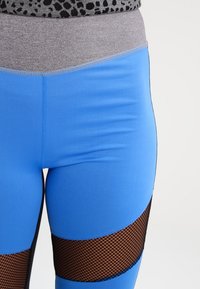 Niebieskie legginsy z szarym pasem, gładkiego materiału, z czarnymi siateczkowymi wycięciami na wysokości ud, tworząc efekt warstwowy.