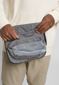 Sacchetto di tessuto grigio con tasca anteriore in rete, chiusura con zip e bordi neri; tenuto saldamente in mano su uno sfondo di abbigliamento chiaro.