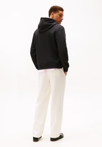 Felpa nera con cappuccio dalla texture liscia, abbinata a pantaloni color crema, larghi e comodi, e scarpe nere, vista da dietro.