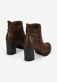 Botas de tobillo de cuero marrón con tacones gruesos apilados, cremalleras laterales, correa decorativa con acentos metálicos y lengüetas en la parte trasera.