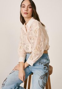 Joven sentada en un taburete, con una blusa transparente crema con bordados florales y unos vaqueros azul claro con diseños florales oscuros.