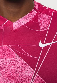 Różowa sportowa koszulka Nike z teksturowanym wzorem i dekoltem w serek. Na przodzie znajduje się białe logo Swoosh. Gładka tkanina z geometrycznymi wzorami.