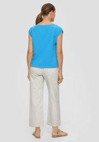 Blaues Kurzarm-Oberteil mit lockerer Passform, kombiniert mit hellgrauen weiten Hosen und braunen Riemchensandalen, von hinten gesehen.