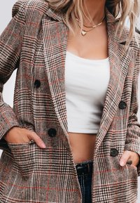 Blazer à carreaux marron avec des boutons noirs. Il présente un col cranté, des poches et un mélange de motifs pied-de-poule et à carreaux avec des accents orange.