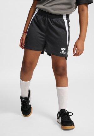 Kind trägt schwarze Sportshorts mit weißen Akzenten, weiße Crew-Socken und schwarze Sportschuhe mit Gummisohlen, steht vor einem schlichten Hintergrund.
