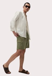 Biała lniana koszula na guziki, beżowy top na ramiączkach, oliwkowo-zielone szorty bojówki, brązowe sandały i czarne okulary przeciwsłoneczne na męskim modelu.