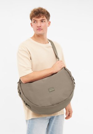 Borsa a spalla in tela verde oliva con forma arrotondata, tracolla regolabile e patch con logo. Indossata in stile a tracolla.
