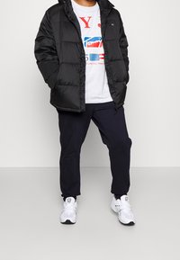Giacca imbottita nera con cappuccio, indossata sopra una maglietta grafica grigia, abbinata a pantaloni scuri e sneakers bianche. Texture imbottita, design casual.