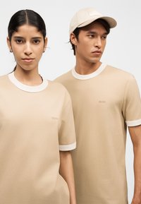 Νεαρή γυναίκα και άνδρας φοράνε μπεζ T-shirts BOSS με άσπρη λεπτομέρεια· ο άνδρας φοράει επίσης μπεζ καπέλο που ταιριάζει, και οι δύο κοιτάνε εμπρός σε λευκό φόντο.