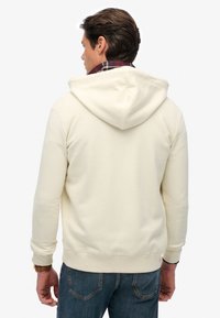 Helle beige Kapuzenjacke mit Kordelzug und aus weichem Stoff, mit gerippten Bündchen und Saum. Getragen über einem rot karierten Hemd.