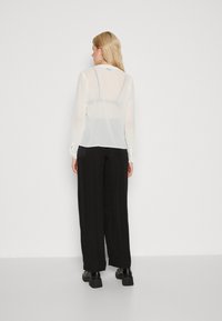 Blusa bianca trasparente a maniche lunghe con una texture liscia, dotata di colletto e polsini, abbinata a pantaloni neri a gamba larga e scarpe nere chunky.