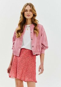Różowa kurtka z haftem i detalami guzików, zestawiona z zwiewną koralową spódnicą w kwiatowy wzór, noszona na białej koszuli.