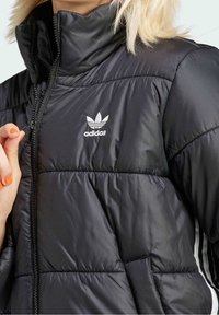 Svart pufferjacka med hög krage, dragkedjestängning, vit Adidas-logga och quiltad struktur med sidofickor.