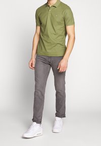 Polo en coton vert à col chemise, associé à un jean gris et des baskets blanches. Le polo présente un petit logo.