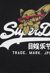 Černá mikina s kapucí s zlatým kresleným psem, který má červené sluneční brýle a dlouhý jazyk, vedle bíleho textu, který říká "SuperD."
