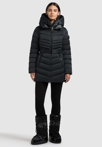 Sort pufferjakke med aftagelig hætte, bælte i taljen, horisontalt quiltet design og lynlåslommer, parret med sorte vinterstøvler.