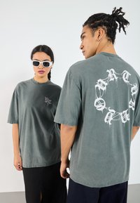 To personer har oversized, afdæmpede grønne T-shirts lavet af blødt bomuld. T-shirtsene har hvide grafiske designs på forsiden og bagsiden.