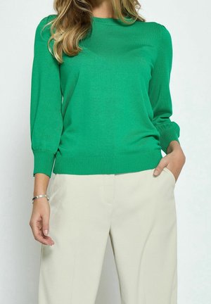 Frau trägt einen leuchtend grünen Pullover mit langen Ärmeln und hellbeige Hose, eine Hand in der Tasche und ein silbernes Armband am Handgelenk.