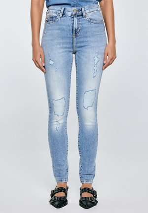 hele sinised rebitud kitsad teksad, mida kannab püsti olev inimene, ühendatud mustade teravate kingadega, millel on pandla rihmad ja silmad.