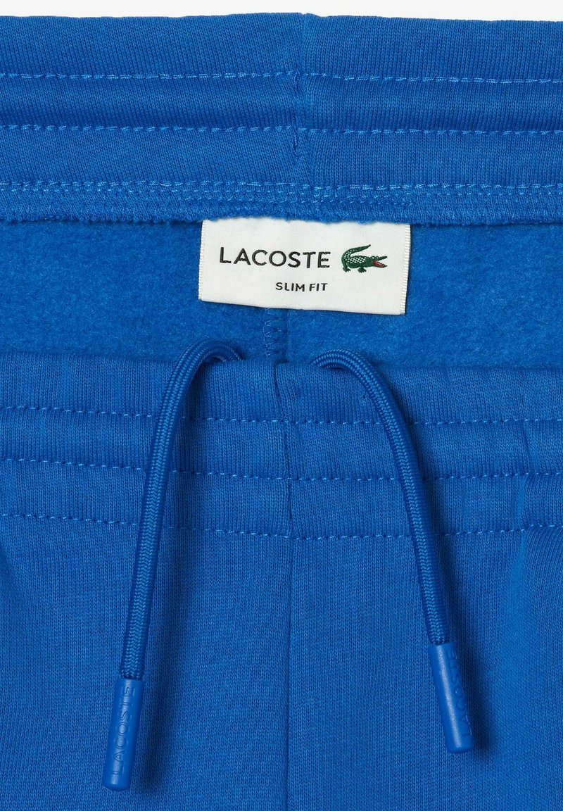Niebieskie dopasowane spodnie z elastycznym paskiem i przeszytymi sznurkami, z białą metką Lacoste i zielonym logo krokodyla.
