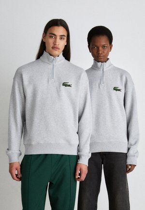 Sudaderas grises con cremallera, con puños y dobladillos acanalados, que presentan un logotipo de cocodrilo bordado en verde en la parte izquierda del pecho.