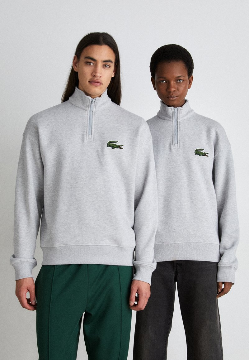 Pelēki zip-up sporta krekli ar ribotām piedurknēm un apakšmalām, ar zaļu izšūtu krokodila logo kreisajā krūtīs.